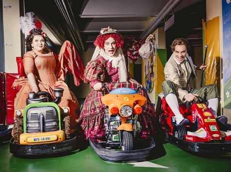 Drei Schauspieler*innen fahren auf Fahrzeugen, wie man sie bei Kinderkarussellen findet und schauen motiviert in die Kamera.