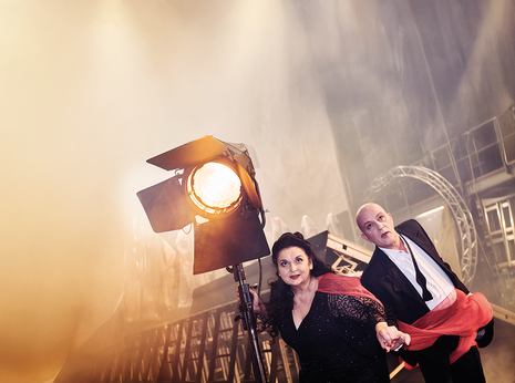 Romelia Lichtenstein und Gerd Vogel halten sich an den Händen. Romelia Lichtenstein hält sich an einem leuchtenden Scheinwerfer. Die beiden gucken konzentriert gerade aus.