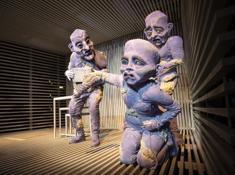 Nils Dreschke, Luise Friederike Hennig und Franziska Dittrich stehen in surrealen, violetten Ganzkörperkostümen mit großen, ausdrucksstarken Masken auf einer Bühne aus Holzlatten. Luise Friederike Hennig kniet mit ausgestrecktem Arm im Vordergrund, während die anderen beiden dahinter stehen und sie angucken.