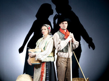Sybille Kreß und Marian Kindermann stehen in traditioneller Robe zwischen Besen und Getreidesäcken. Im Hintergrund sind Schatten von zwei Personen, die sich küssen.