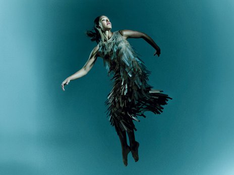 Eine Frau in einem kunstvollen, fransigen Kleid scheint schwerelos in der Luft zu schweben, vor einem weichen, blauen Farbverlauf als Hintergrund.