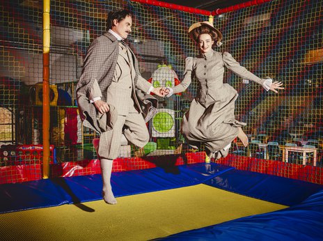 Robert Sellier und Yulia Sokolik, Opernsänger*innen, springen lachend in historischen Kostümen auf einem Trampolin in einem bunten Indoor-Spielplatz.