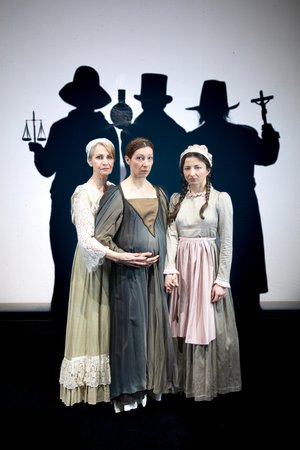 Elke Richter hält den schwangeren Bauch von Nicoline Schubert, während diese Kinga Schmidts Hand hält. Im Hintergrund sind drei Personen mit Hüten, welche Waage, Reagenzglas und Kruzifix in den Händen halten. © Julia Fenske