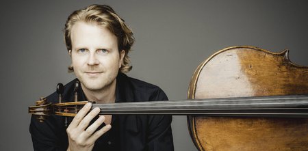 Cellist Julian Steckel lächelt leicht in die Kamera und hält ein Violoncello, dass quer zu ihm liegt. © Arnaud Ele