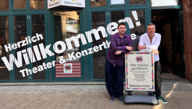 Zwei Männer lehnen an einem Aufsteller mit der Aufschrift „Wir haben geöffnet!“. Links steht Ballettdirektor Michal Sedláček in einem lila gemusterten Hemd, rechts Opernintendant Walter Sutcliffe in einem weißen Hemd mit hochgekrempelten Ärmeln. Hinter ihnen begrüßt ein großes Schaufensterdesign mit „Herzlich Willkommen! Theater & Konzertkasse“ die Besucher*innen.
