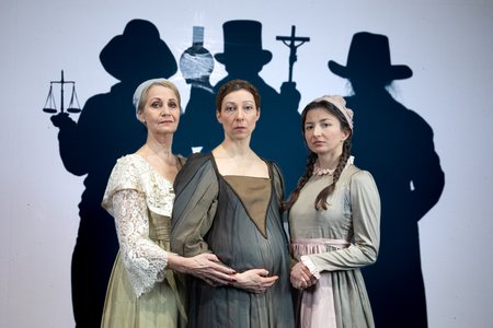 Elke Richter hält den schwangeren Bauch von Nicoline Schubert, während diese Kinga Schmidts Hand hält. Im Hintergrund sind drei Personen mit Hüten, welche Waage, Reagenzglas und Kruzifix in den Händen halten. © Julia Fenske